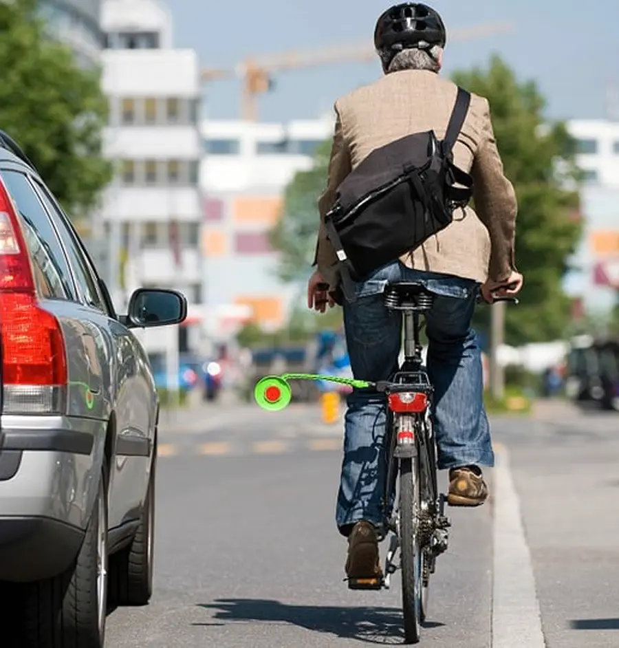 Ein Radfahrer mit einer am Gepäckträger montierten Abstandskelle wird trotzdem von einem Auto mit geringem Abstand überholt. Ein Radfahrer im Stadtverkehr nutzt eine am Gepäckträger montierte Abstandskelle, um überholende Autos auf den nötigen Sicherheitsabstand für Radfahrer hinzuweisen. Ein Auto fährt trotzdem sehr knapp an ihm vorbei. Der Fahrrad Abstandshalter Safety Spacer kann das vermeiden.
