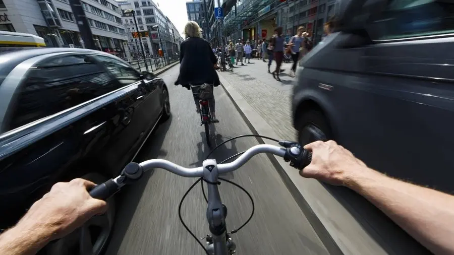 Gefährliche Enge im Stadtverkehr aus der Perspektive eines Radfahrers. Aus der Perspektive eines Radfahrers ist zu sehen, wie er und eine vor ihm fahrende Radfahrerin mit viel zu kleinem Abstand von einem Auto überholt werden. Nach rechts auszuweichen ist wegen der Bordsteinkante und einem parkenden Lieferwagen nicht möglich. Typische Situation, wenn im Stadtverkehr der Sicherheitsabstand für Radfahrer missachtet wird. Der Fahrrad Abstandshalter Safety Spacer kann das vermeiden.
