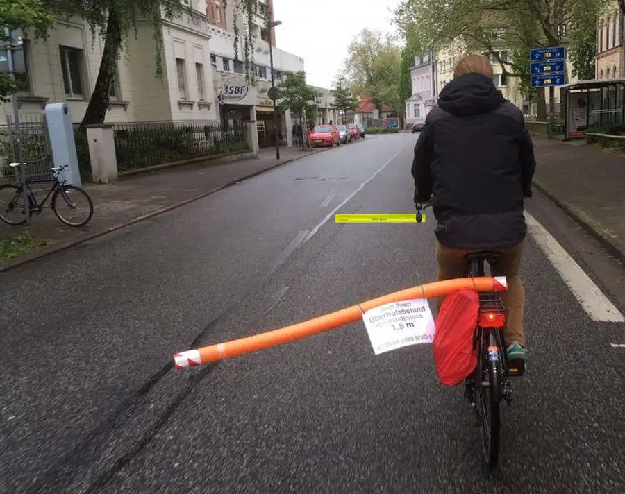 A cyclist uses a DIY spacer made from an orange pool noodle, with a sign demanding the legal 1.5m passing distance, to enforce a safe passing distance for cyclists.
